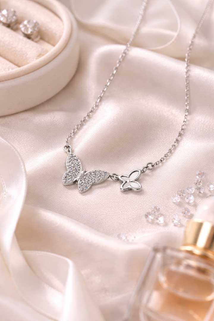 A silver necklace featuring two butterfly charms rests on a silky, light-colored fabric near jewelry and perfume.