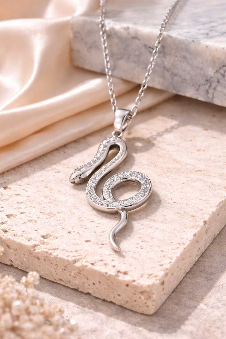 A silver, snake-shaped pendant encrusted with small clear stones, hanging on a matching chain against a stone surface.
