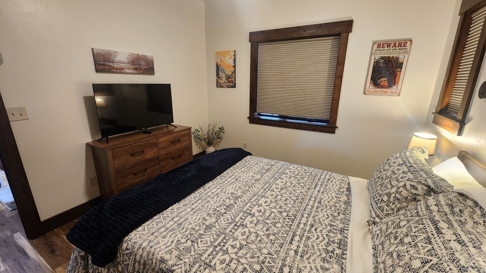 Bedroom with bed, TV, and wooden chest, window with shades, and decorative art on the walls.