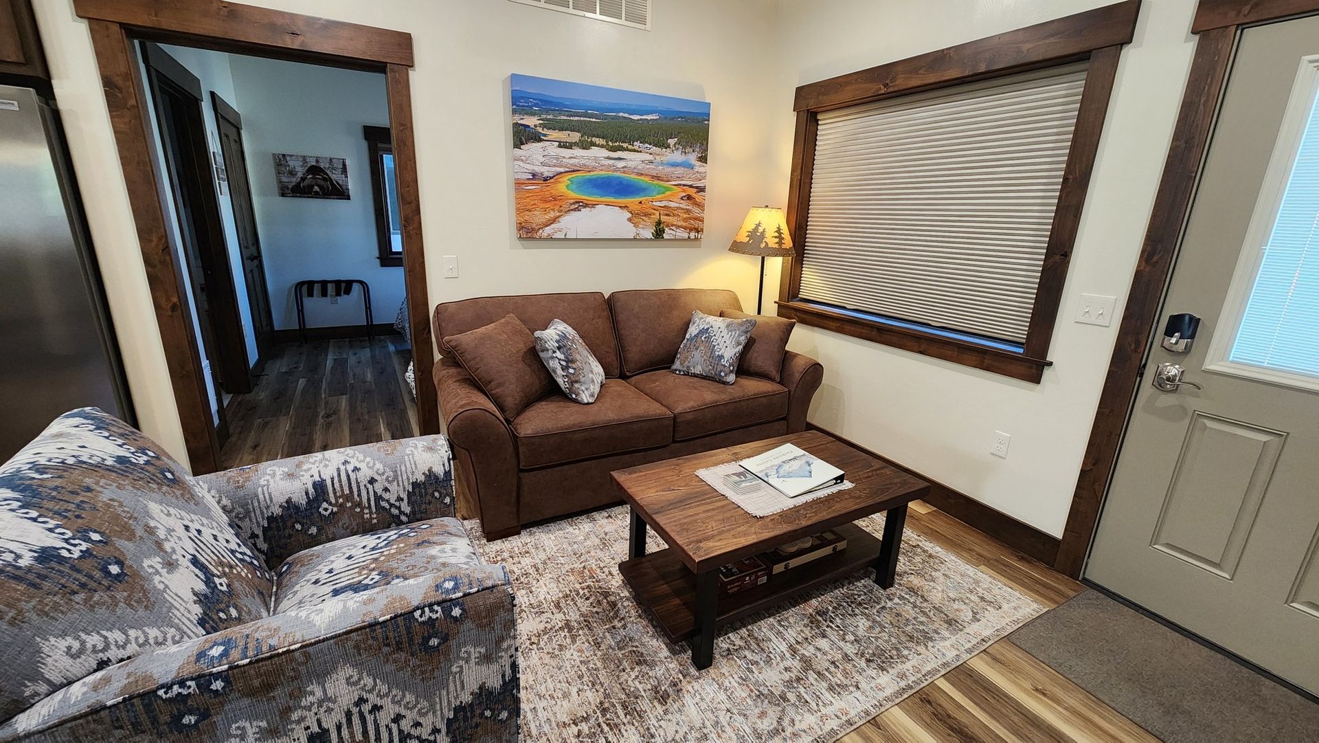 Cozy living room: brown sofa, patterned armchair, wooden table, artwork, rug, and doorway.