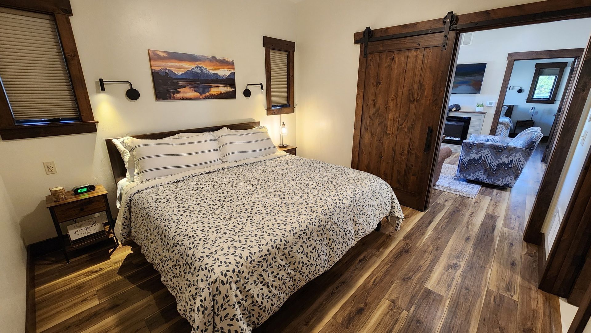 Bedroom with a bed, a barn door leading to a living room, wooden floors, and a mountain landscape painting.
