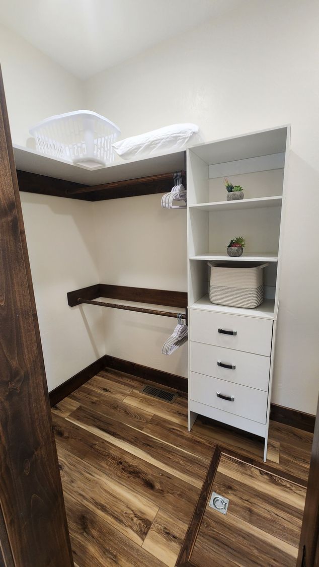 Small walk-in closet with white shelves, drawers, and wood-look floor. Dark trim and a shelf above.