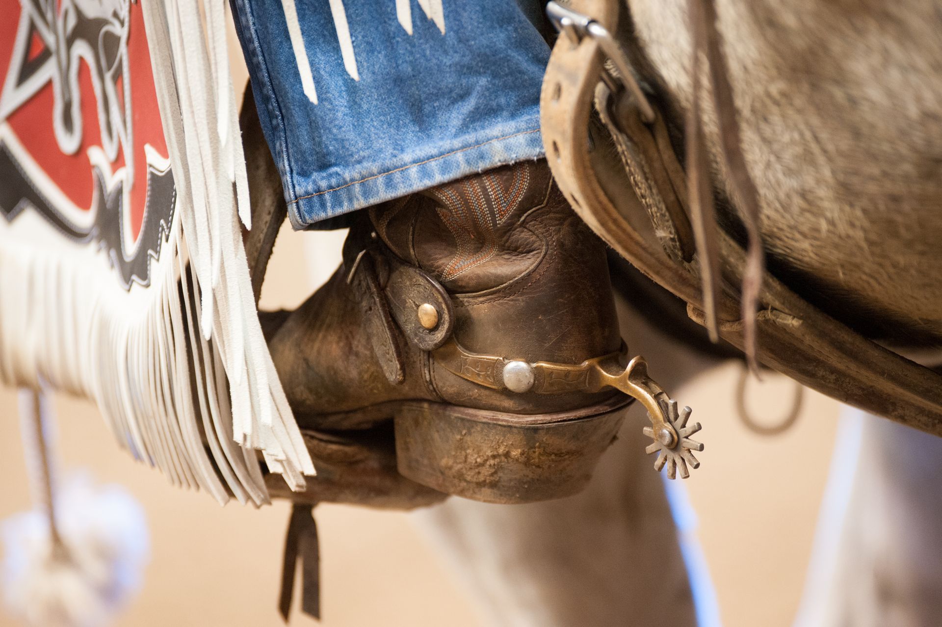 Close-up of a cowboy's boot with spur in a stirrup, riding a horse, fringe of saddle blanket visible.