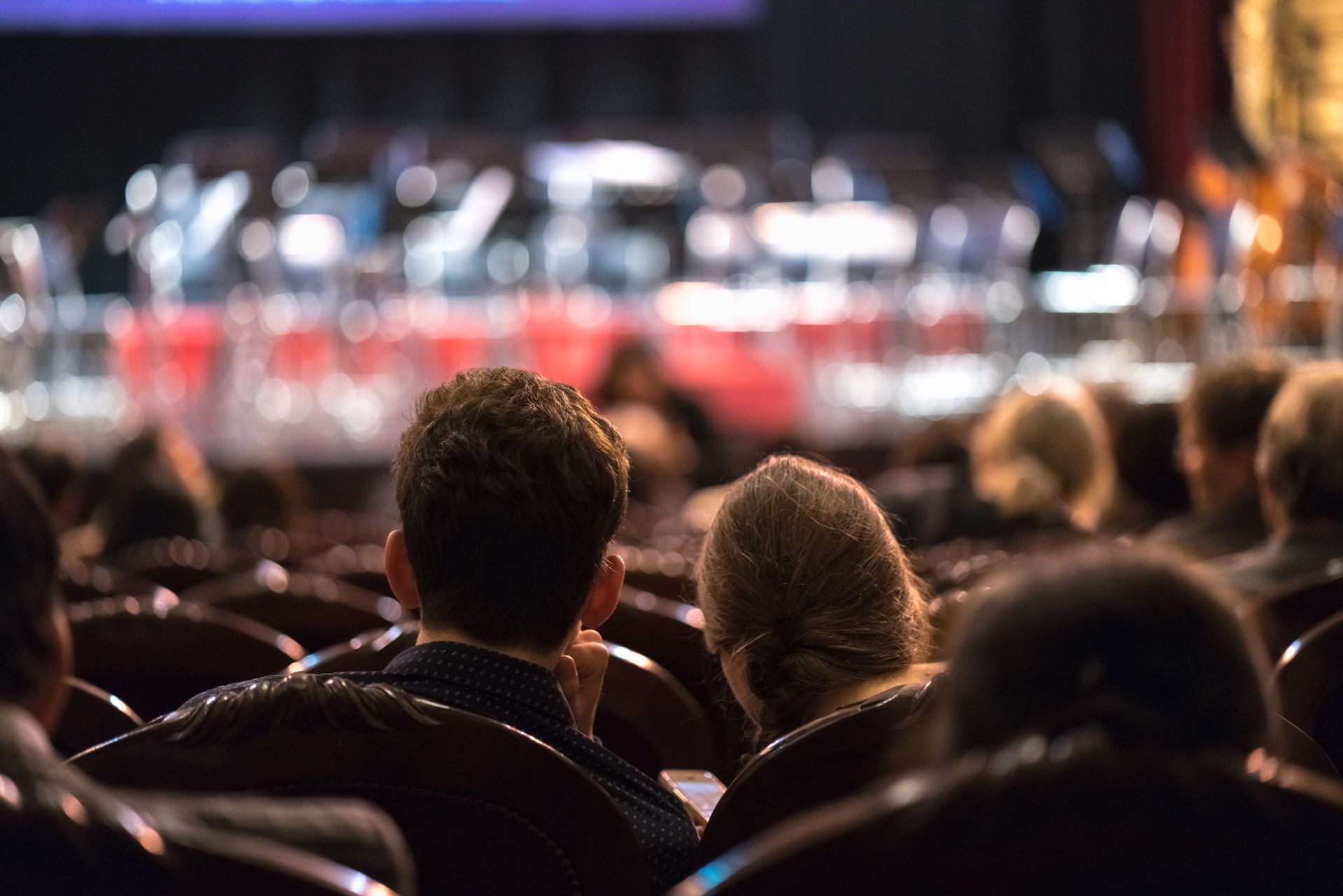 People watching a performance from a theater seat, with the stage out of focus in the background.