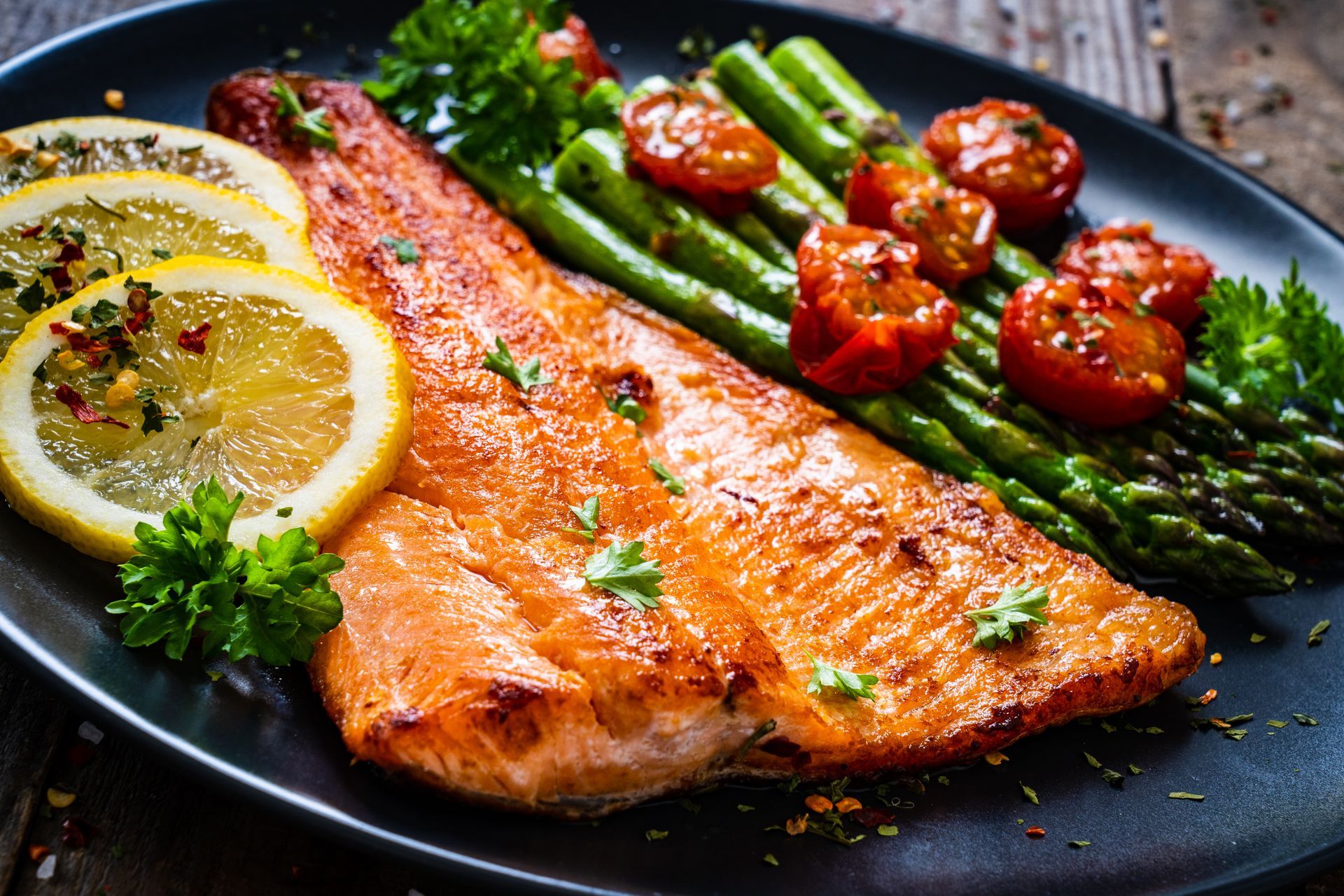 Plate of cooked salmon, asparagus, cherry tomatoes, and lemon slices on a black plate.