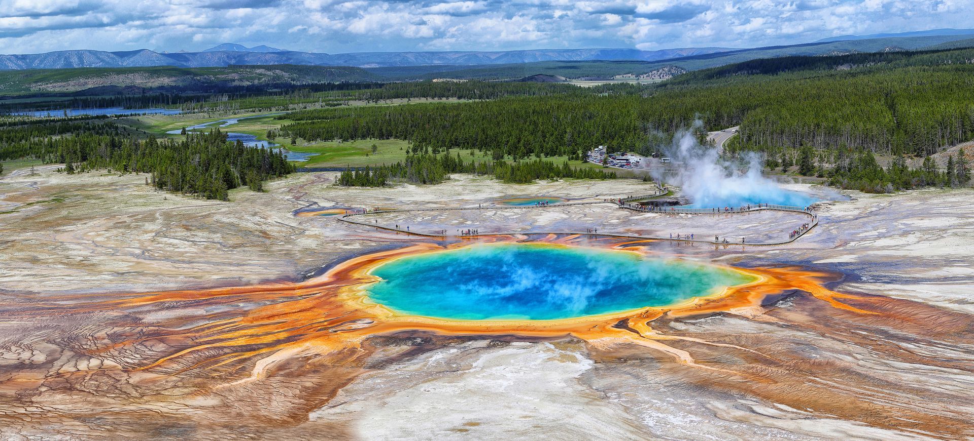 Aerial view of Grand Prismatic Spring in Yellowstone National Park, Wyoming, with colorful hot spring and geyser in background.