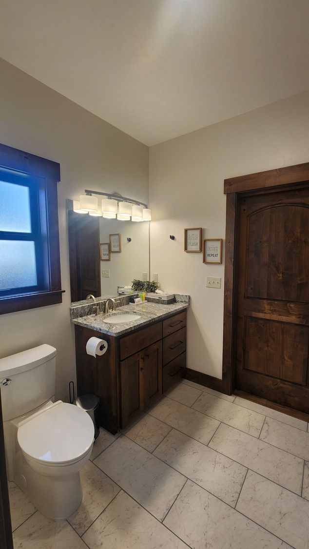 Bathroom with toilet, vanity, and dark wood door. Beige walls, stone floor.