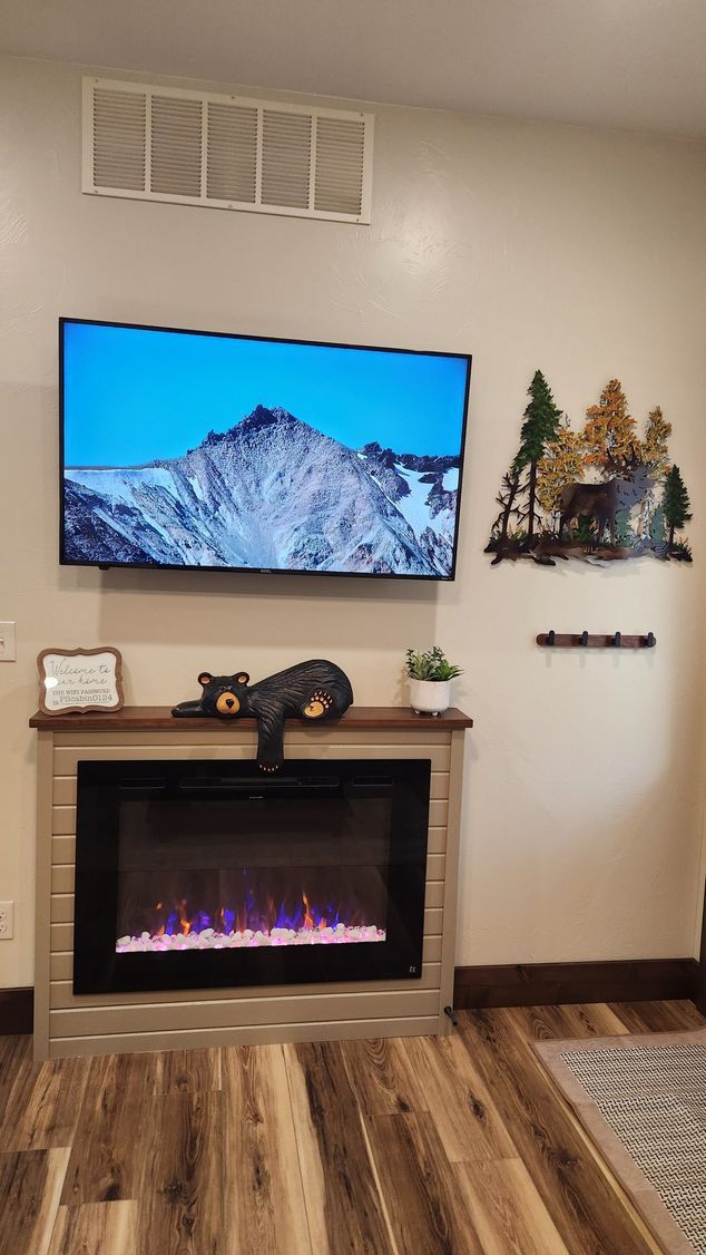 Wall-mounted TV above an electric fireplace, decorated with a bear figurine and metal forest art, on a wood floor.