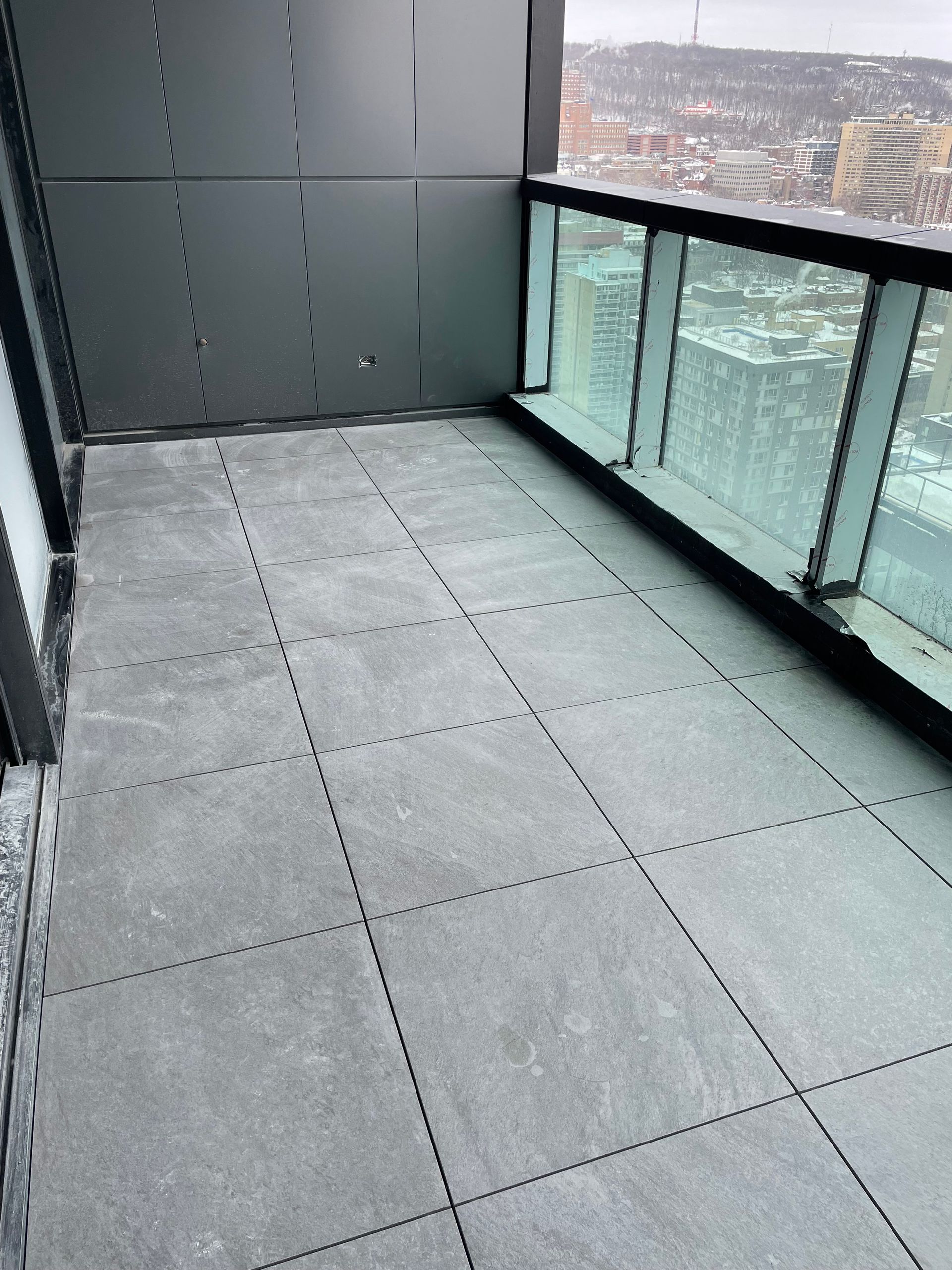 Un balcon en hauteur avec un carrelage gris foncé à carreaux emboîtables, une rambarde en verre et une vue sur une ville enneigée.