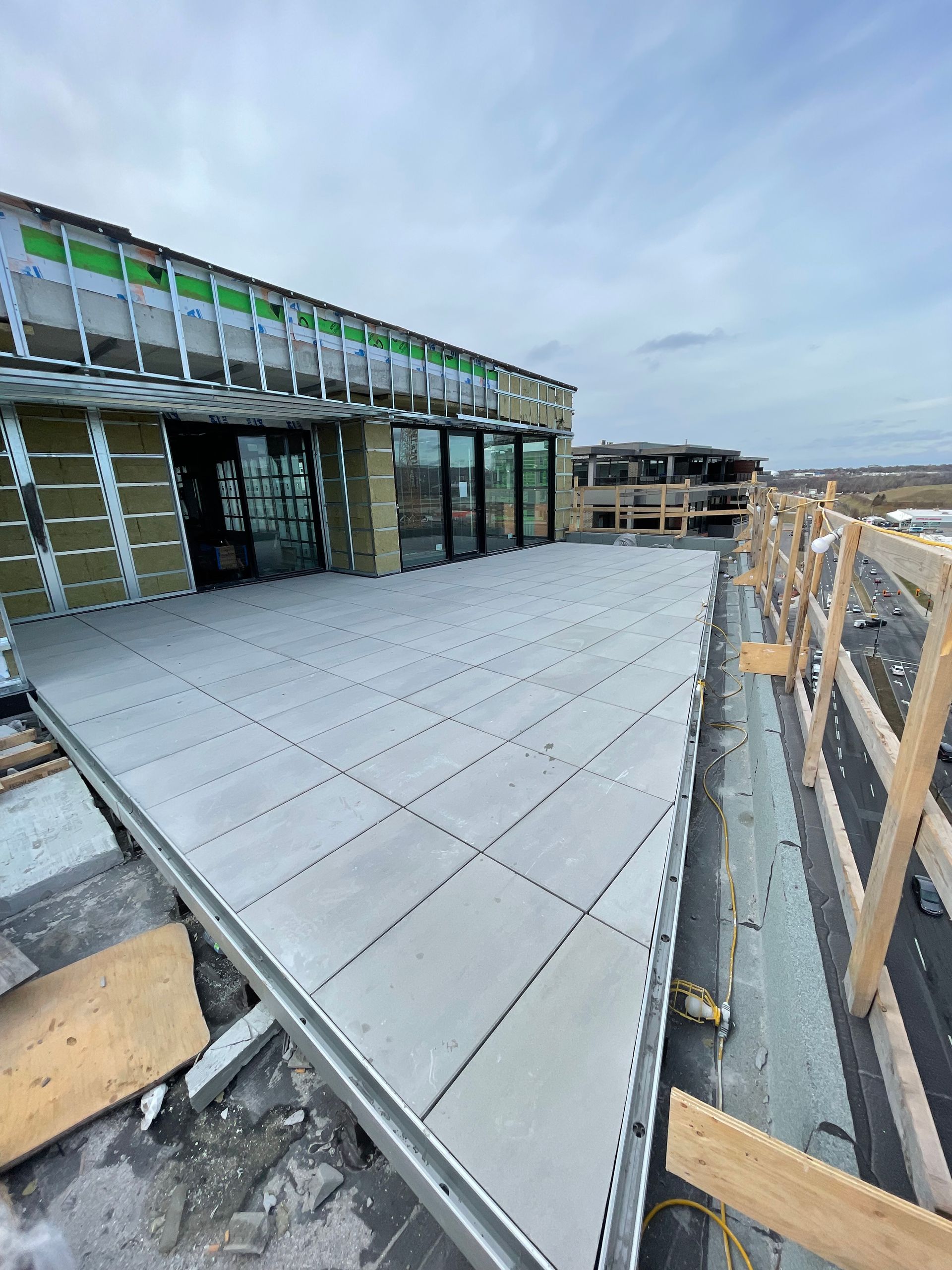 Un chantier de construction sur un toit-terrasse montrant une terrasse en pavés gris nouvellement installée, adjacente à la façade d'un bâtiment avec des portes vitrées.