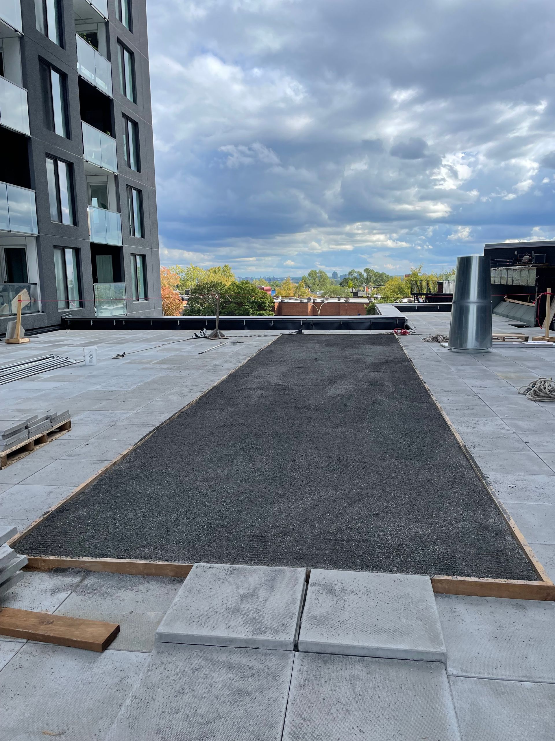 Un rectangle de gravier sombre entouré de dalles de béton gris sur un toit plat à usage industriel, sous un ciel nuageux.