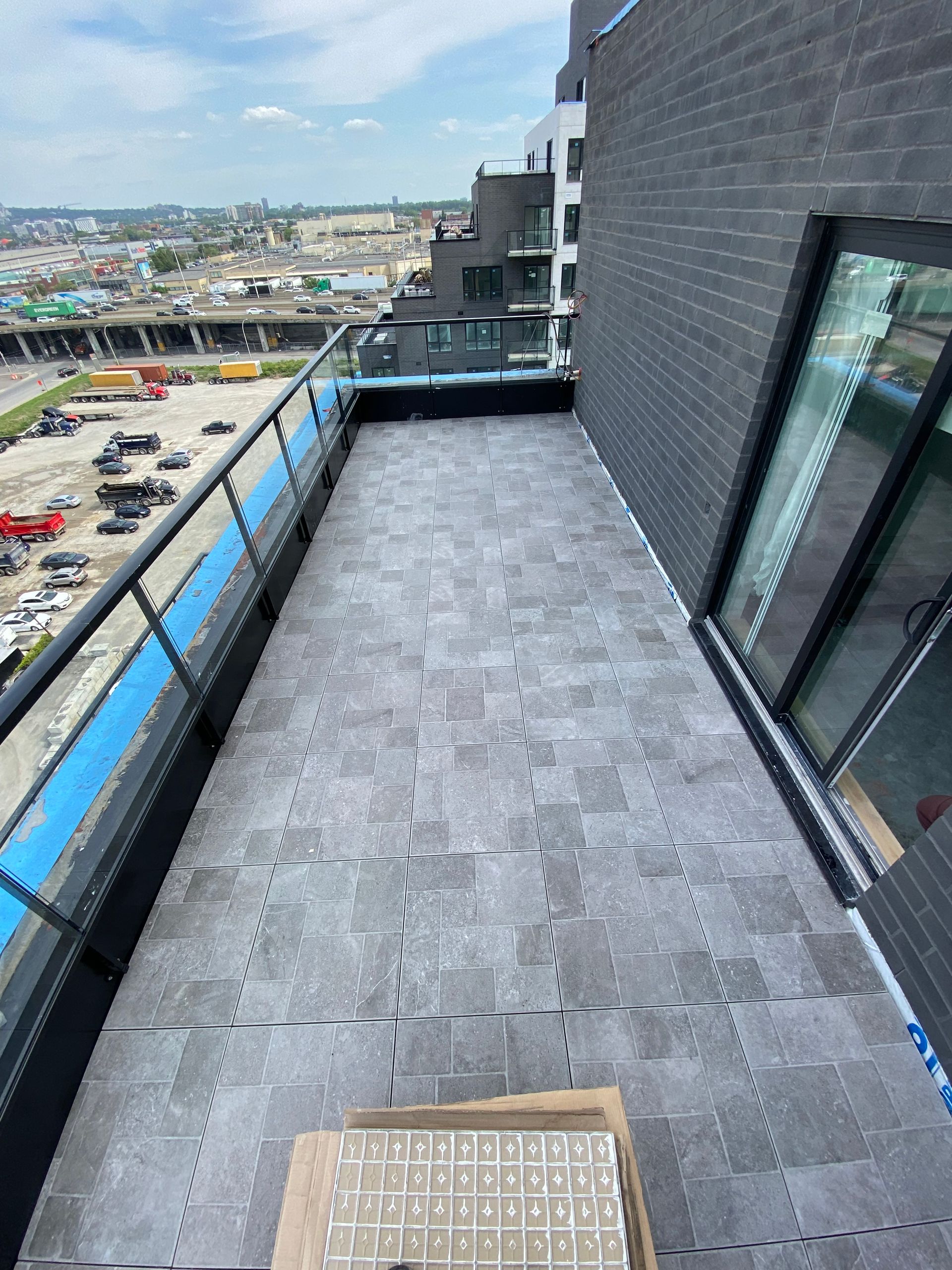 Vue en plongée d'un balcon d'appartement carrelé de gris avec une rambarde métallique, donnant sur un parking et des immeubles de la ville.