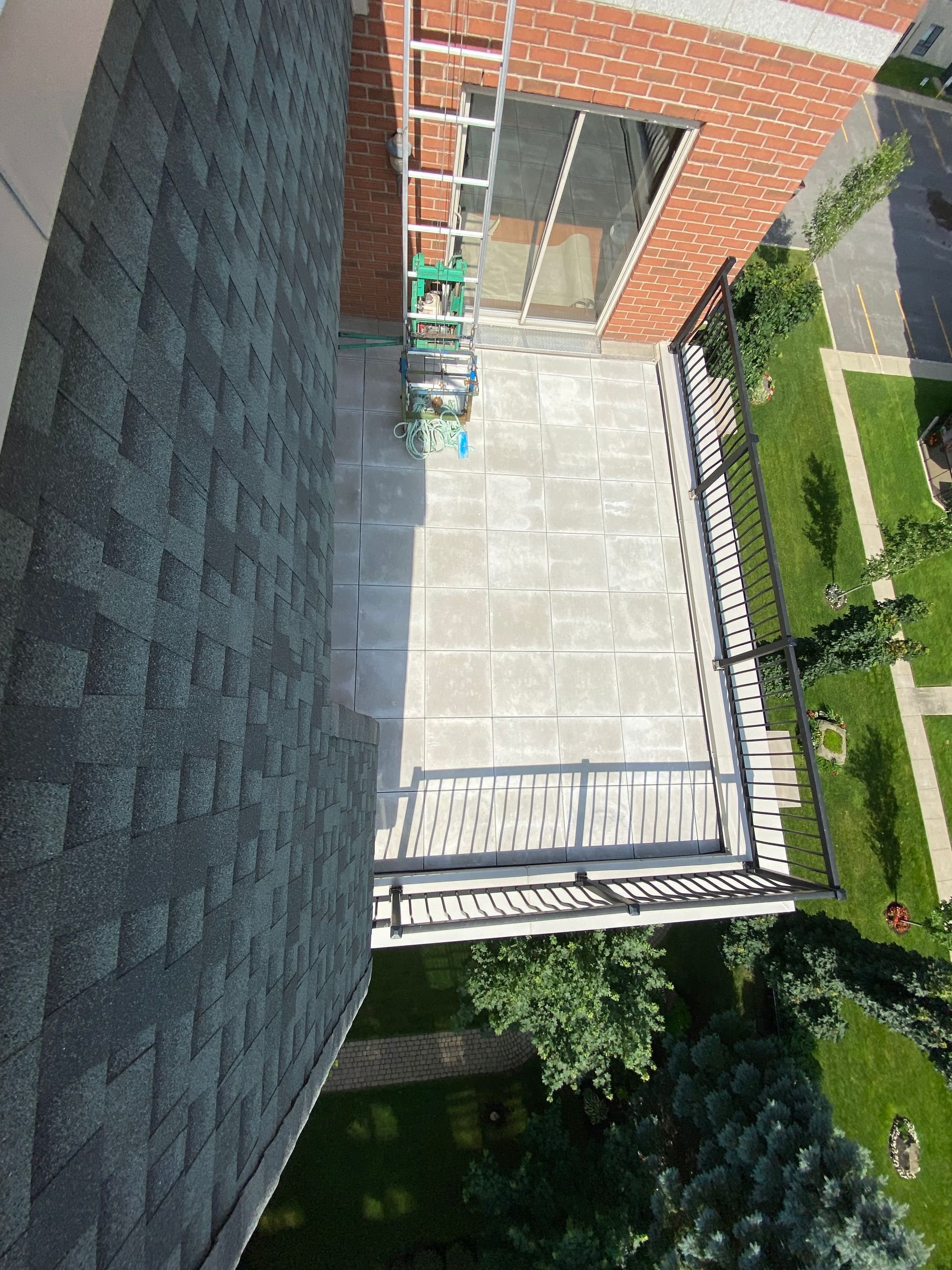 Vue aérienne d'un balcon carrelé de gris avec une rambarde noire, à côté d'un toit en bardeaux et d'un bâtiment en briques.