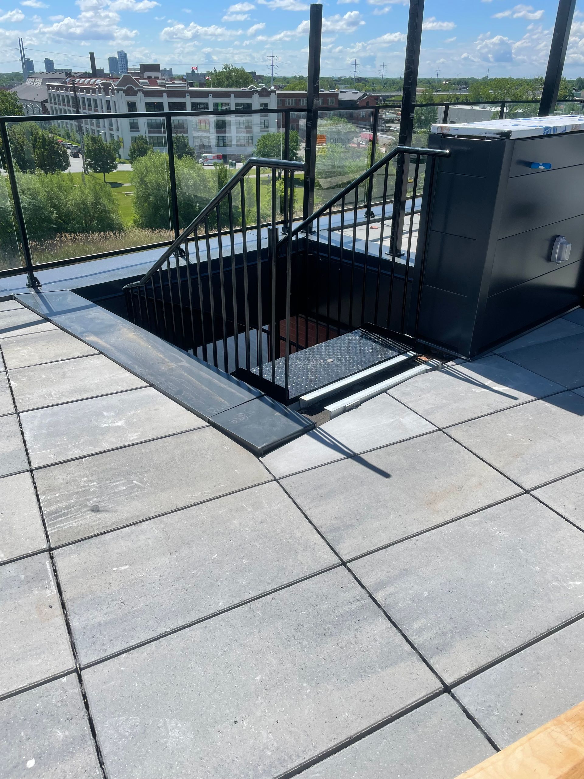Une terrasse sur le toit avec un escalier en métal noir descendant, revêtue de dalles grises et dotée d'une rambarde de sécurité noire.