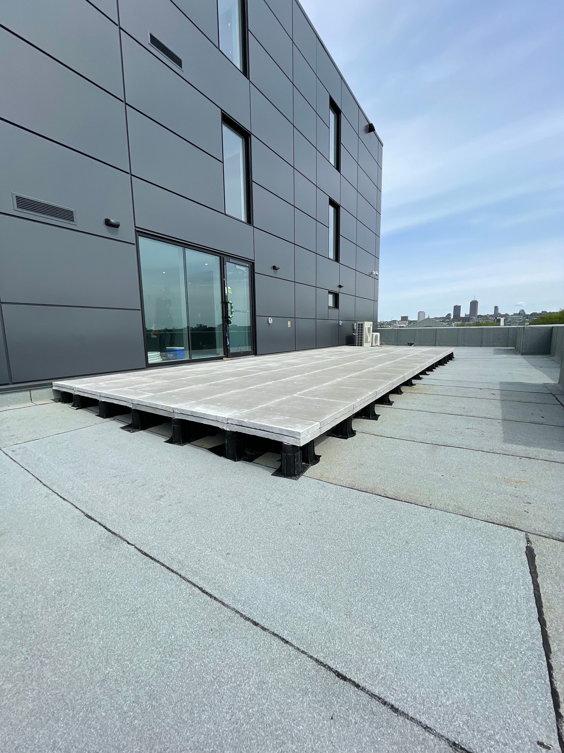 Une plateforme rectangulaire surélevée, composée de panneaux gris clair reposant sur des socles réglables foncés, sur un toit plat gris.