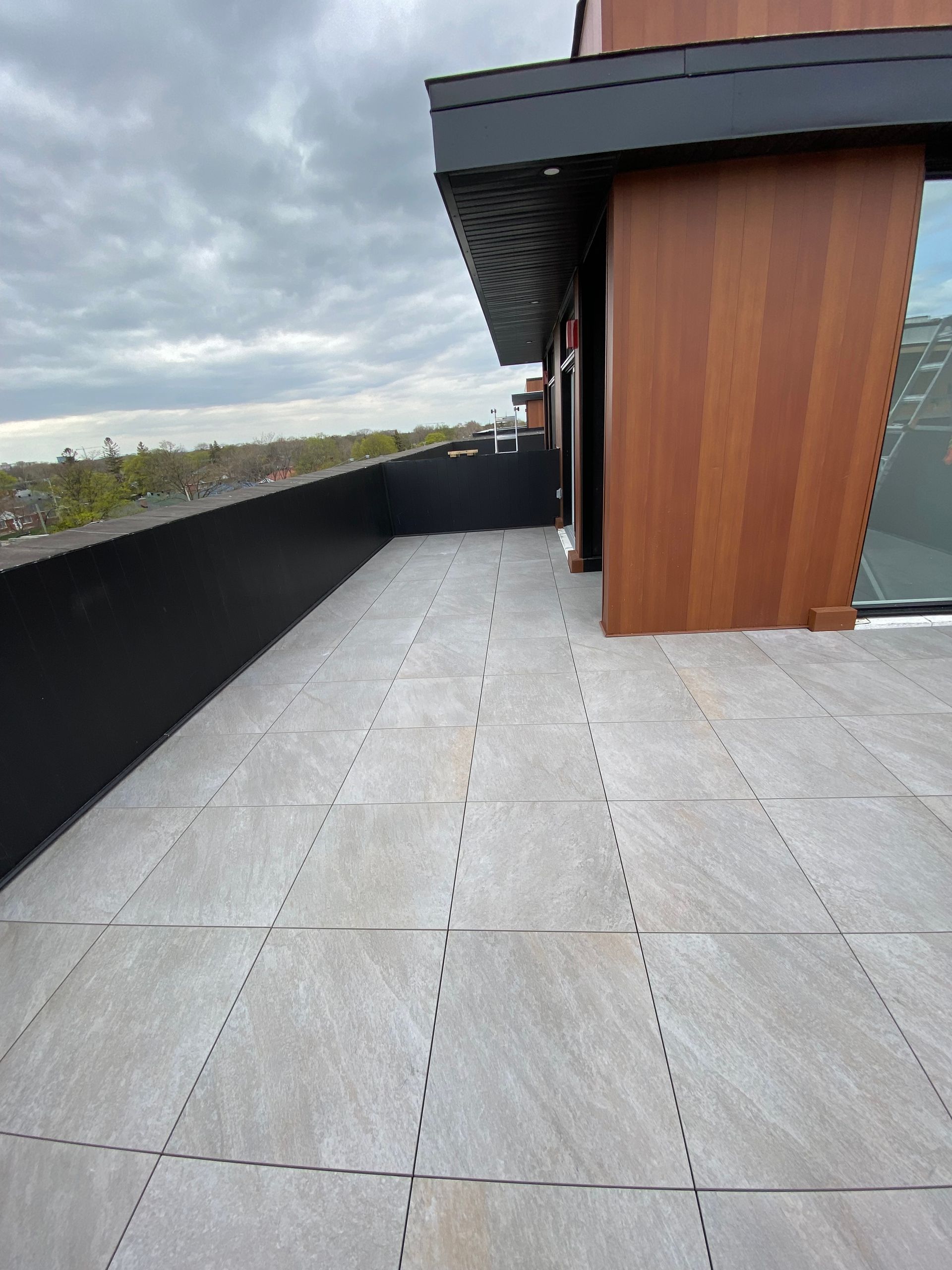 Un balcon moderne sur le toit, avec un sol carrelé en pierre grise, une rambarde en métal noir et une façade en lambris.