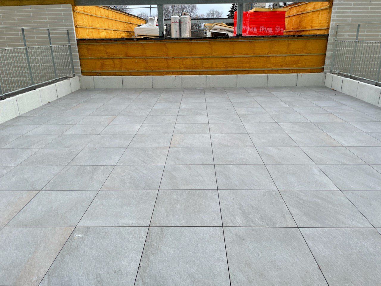 Une terrasse carrelée en dalles de pierre carrées gris clair, entourée d'un muret et d'une clôture métallique.