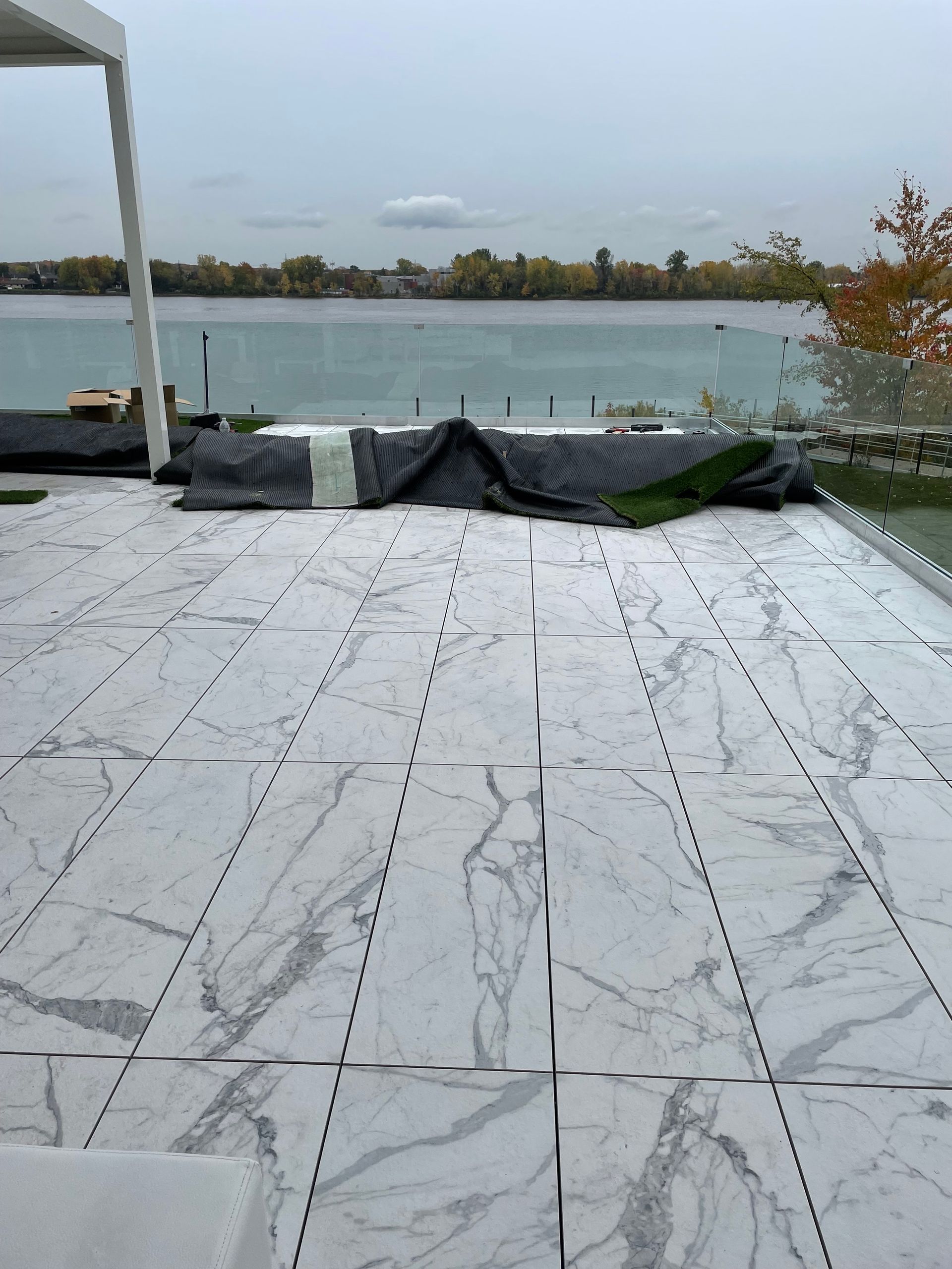De grands carreaux de sol blancs à motifs de marbre ont été installés sur une terrasse extérieure donnant sur une rivière, parmi des débris de construction noirs.