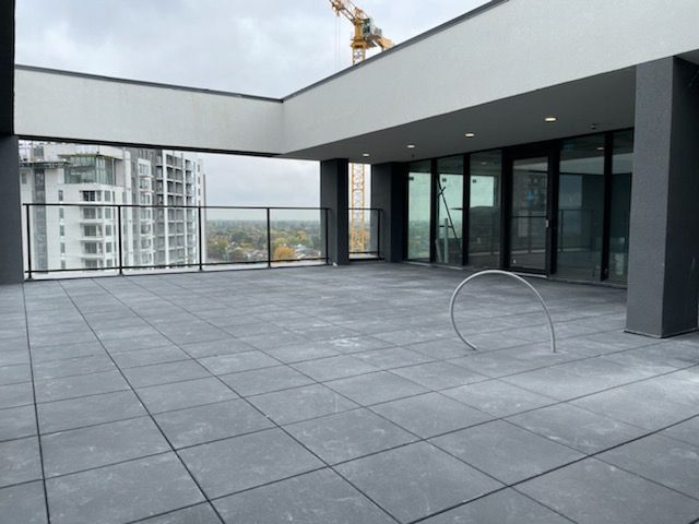 Une terrasse-balcon grise carrelée et vide, avec une balustrade en verre, donnant sur un immeuble d'appartements au loin, sous un ciel nuageux.