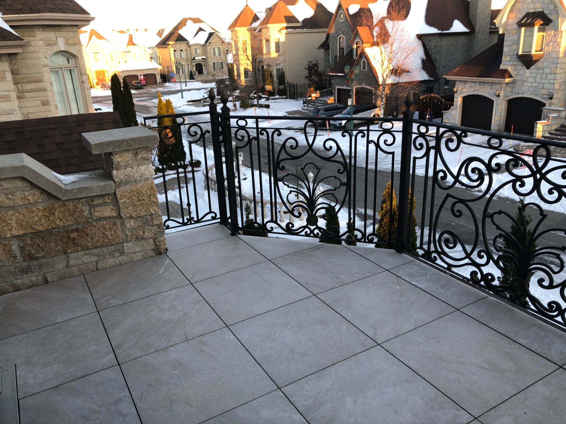 Un balcon en pierre avec une balustrade en fer forgé noir ouvragée donnant sur un quartier résidentiel enneigé.