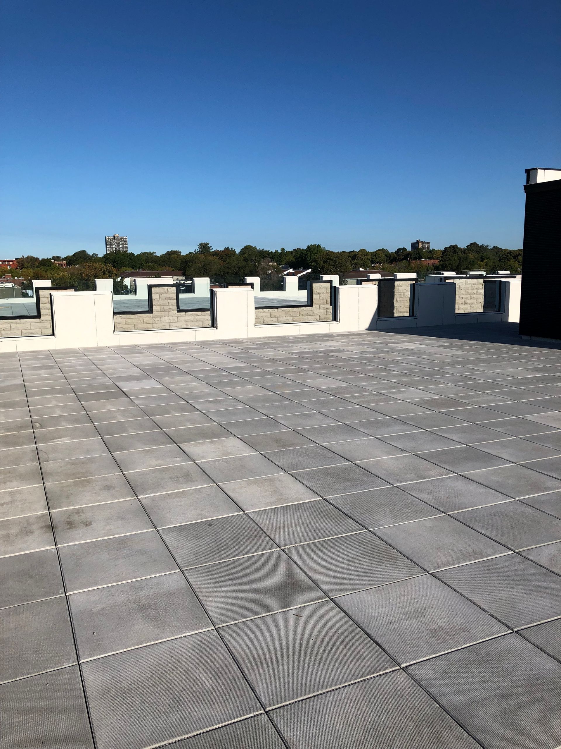 Une terrasse sur le toit, revêtue de dalles carrées grises imbriquées, entourée d'un muret en pierre blanche et grise, sous un ciel bleu.