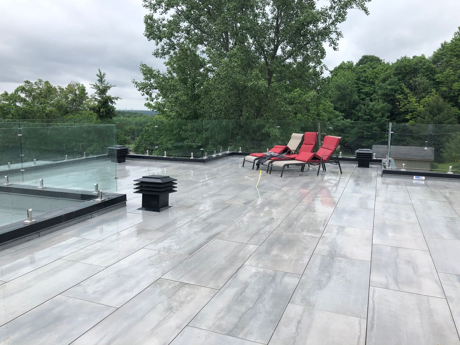 Une terrasse sur le toit avec un sol carrelé gris, trois chaises longues rouges et une balustrade en verre donnant sur un paysage boisé.