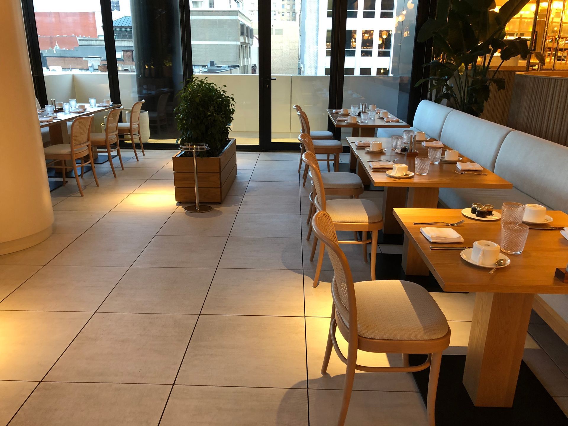 Une terrasse de restaurant lumineuse avec des tables en bois, des chaises claires et de grandes fenêtres donnant sur la ville.