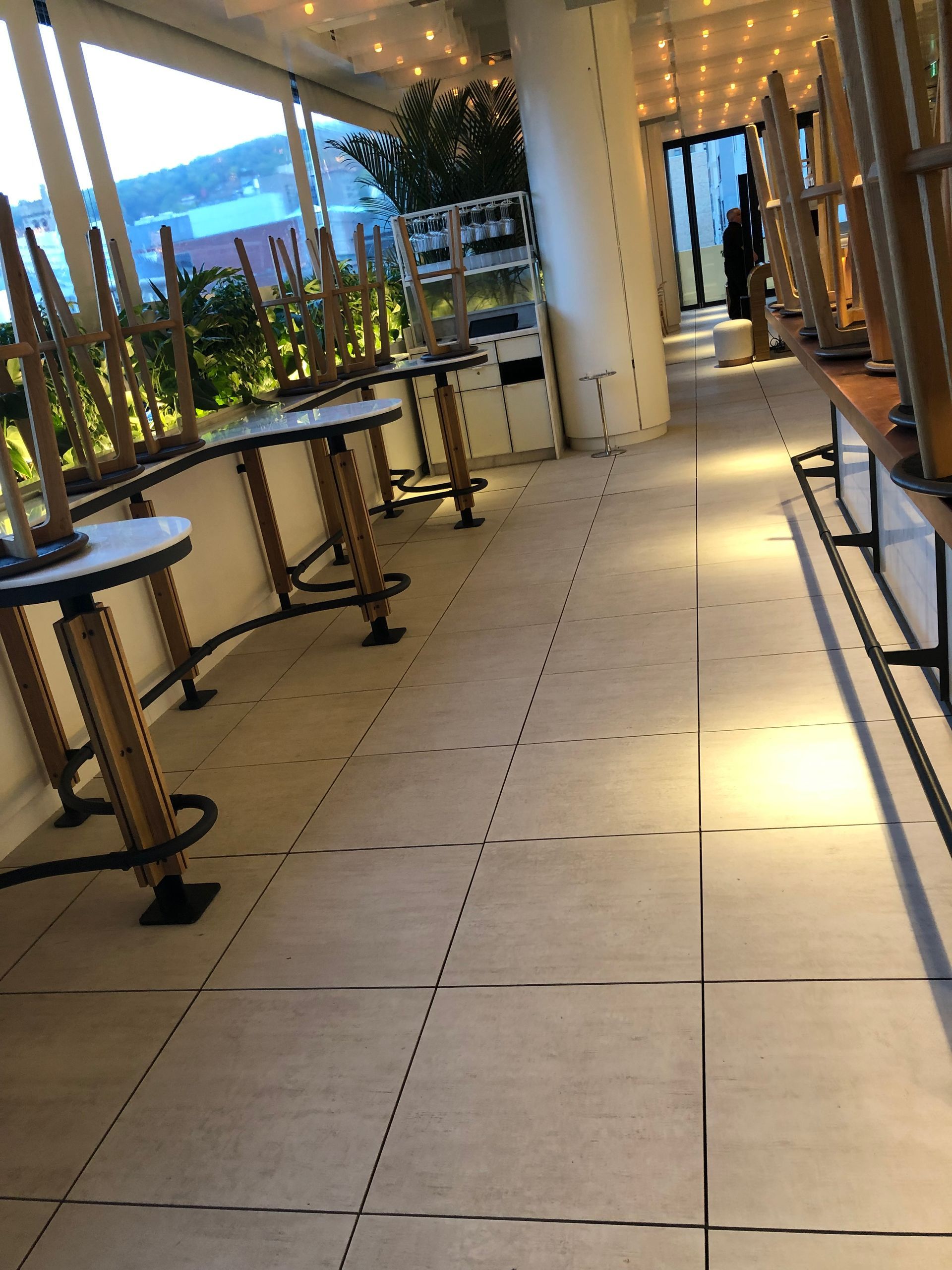 Un bar intérieur avec balcon ou terrasse, des tables et des chaises vides alignées le long d'un mur vitré, et un sol carrelé.