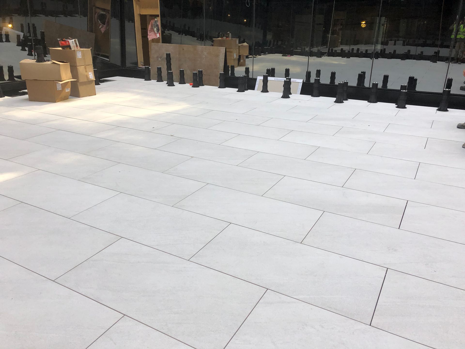 Un chantier de construction montrant la pose de dalles de pavage rectangulaires de couleur claire sur une grille de plots de support noirs.