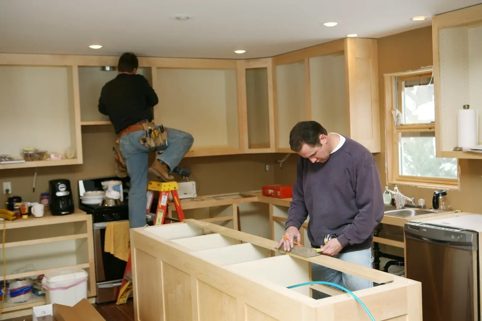 Two people installing kitchen cabinets. One on a ladder, one at the island, working on cabinetry.