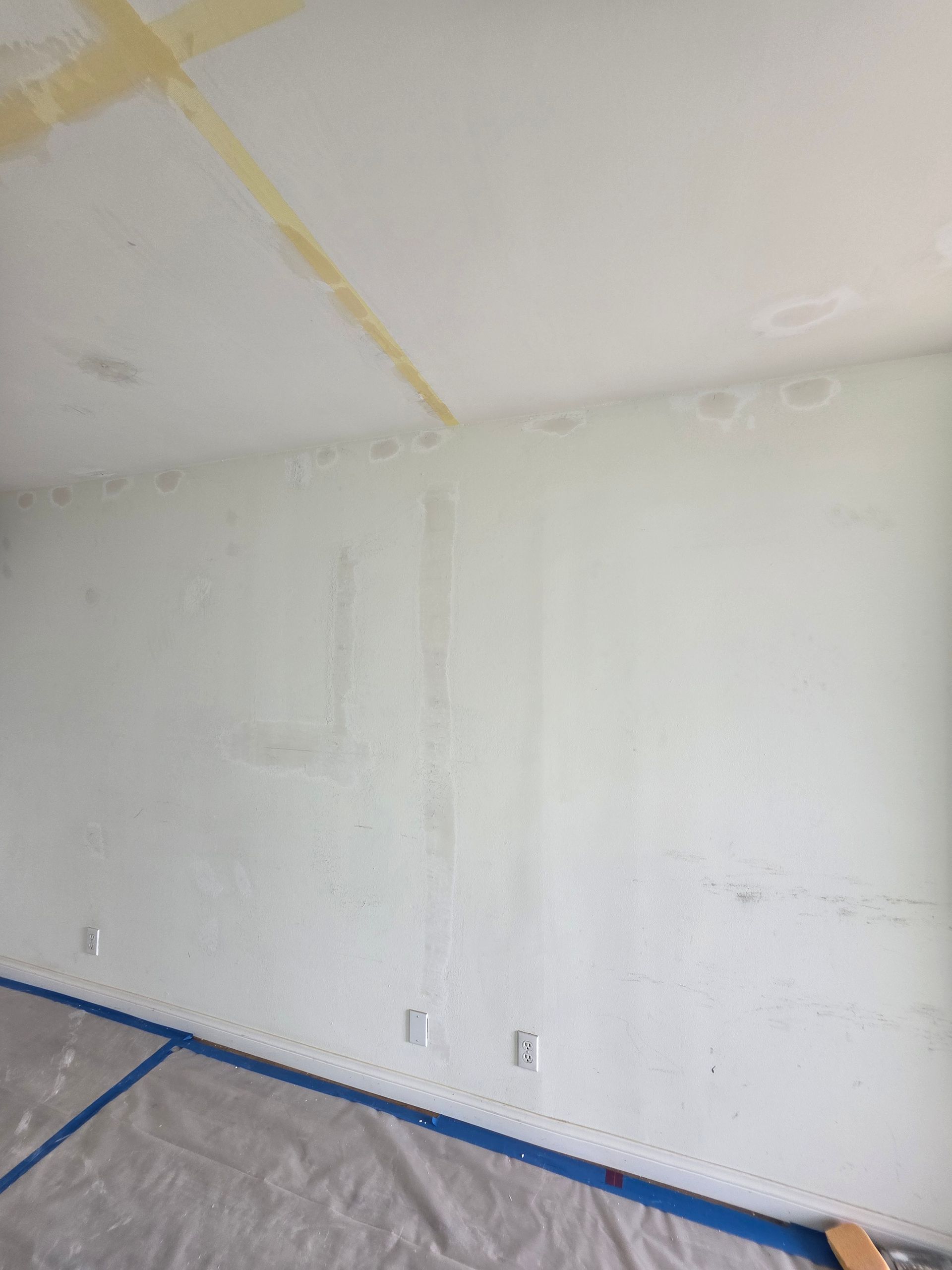 White wall in a room under renovation, with blue tape along the baseboard and plastic sheeting on the floor.