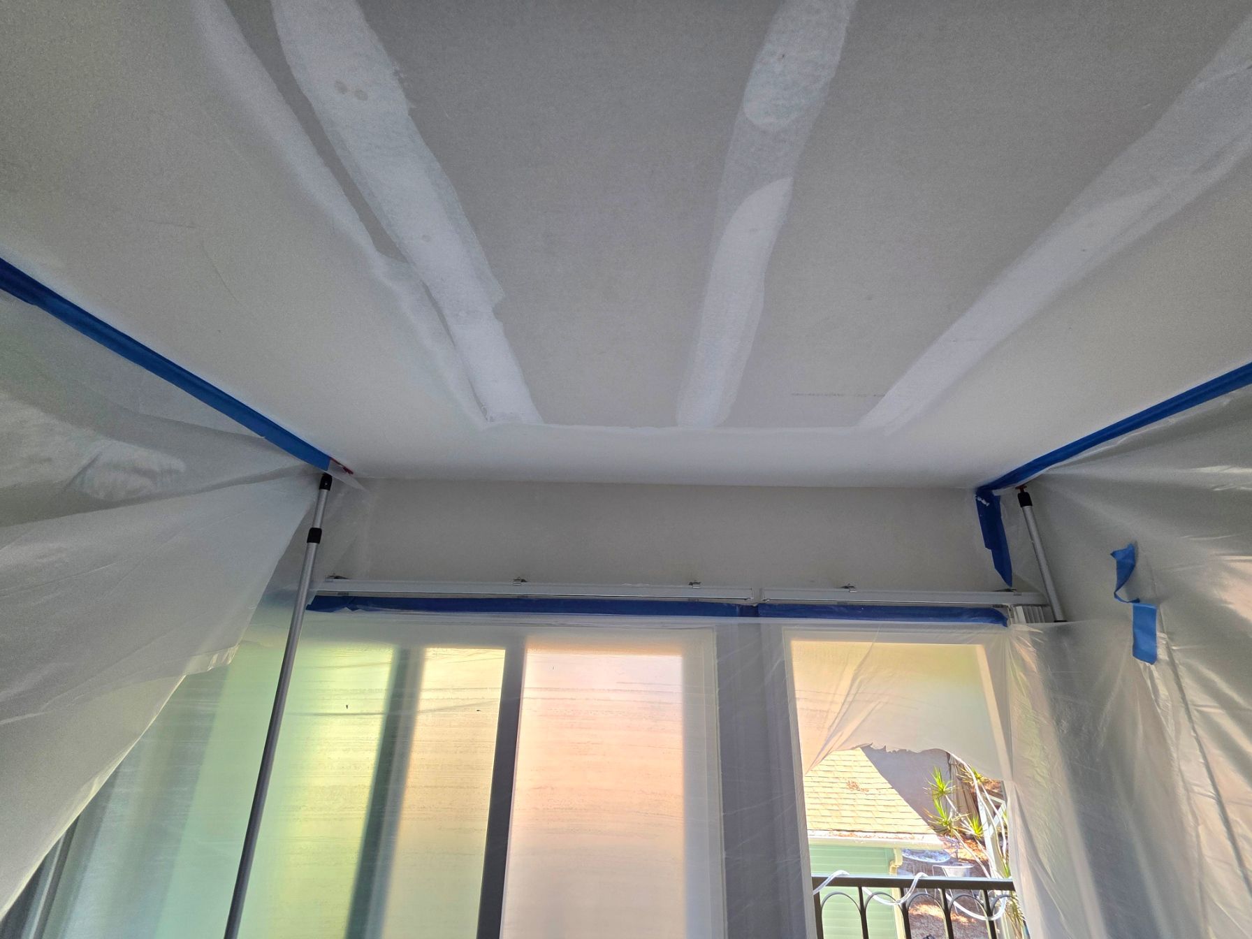 Interior room under renovation with white plastic sheeting, blue tape, and a bright window.