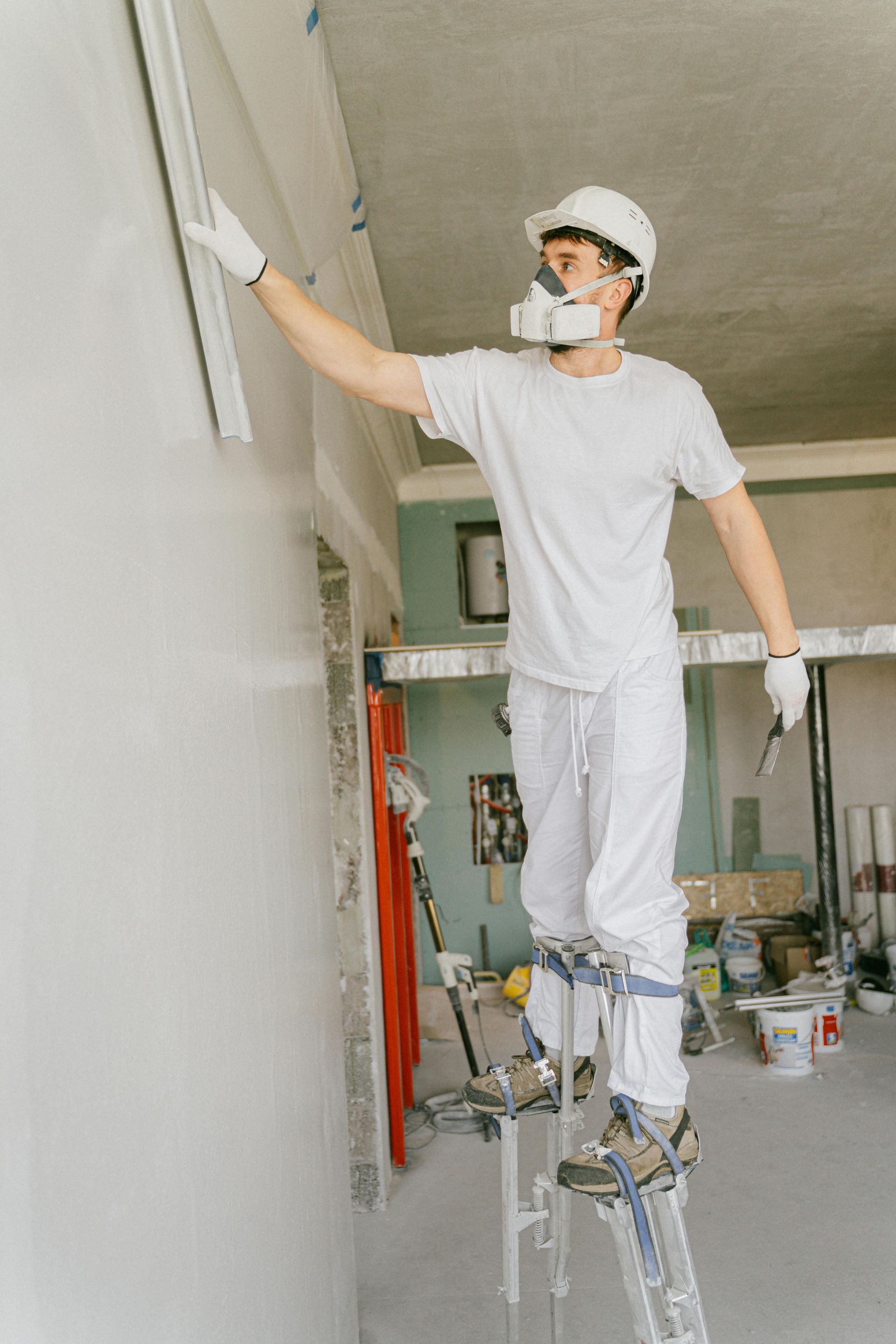 Worker in white protective gear standing on stilts, painting a ceiling indoors.