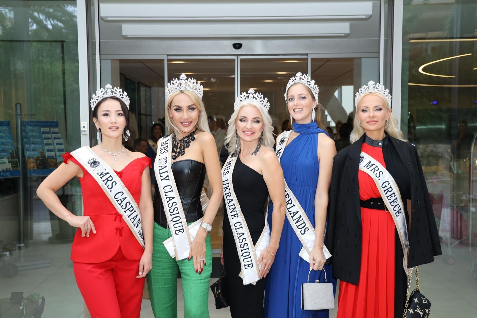 Five women in evening wear pose outside, wearing crowns and sashes.