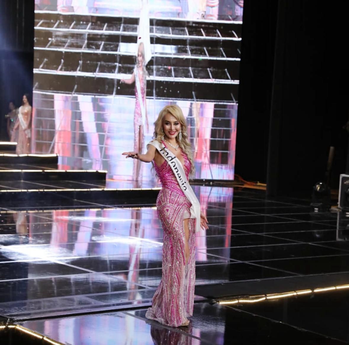 Woman in pink gown with sash poses on stage with mirrored backdrop.