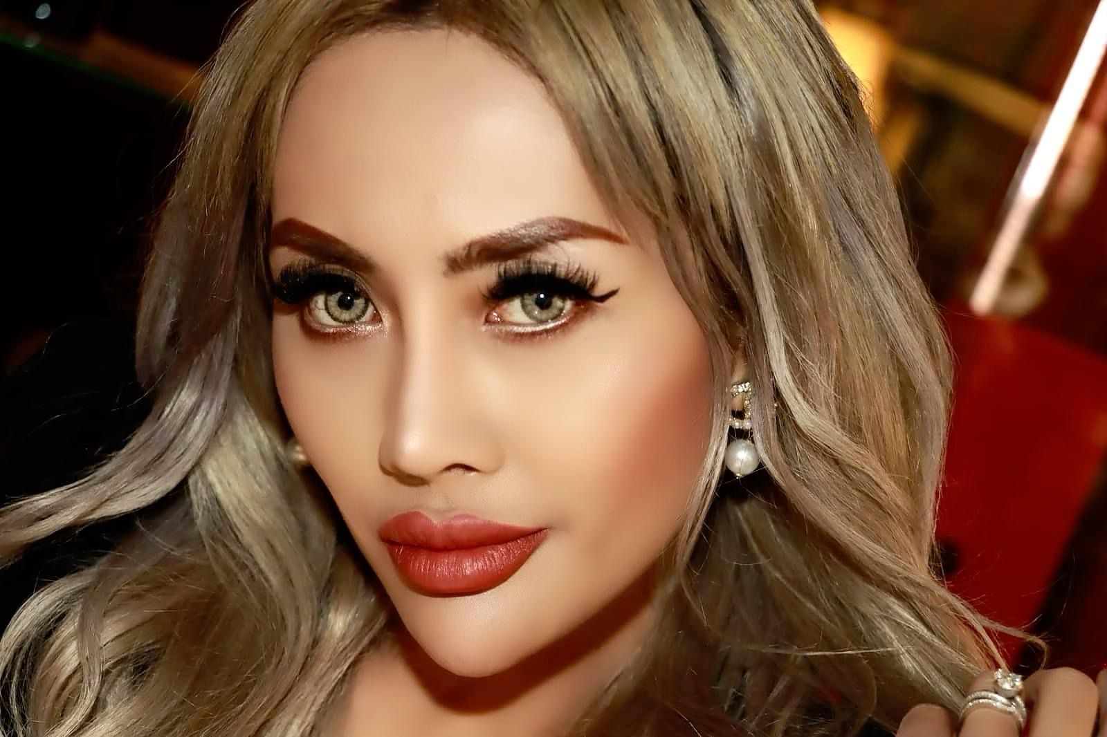 Blonde woman with large eyelashes and red lipstick, wearing pearl earrings, looking towards the camera.