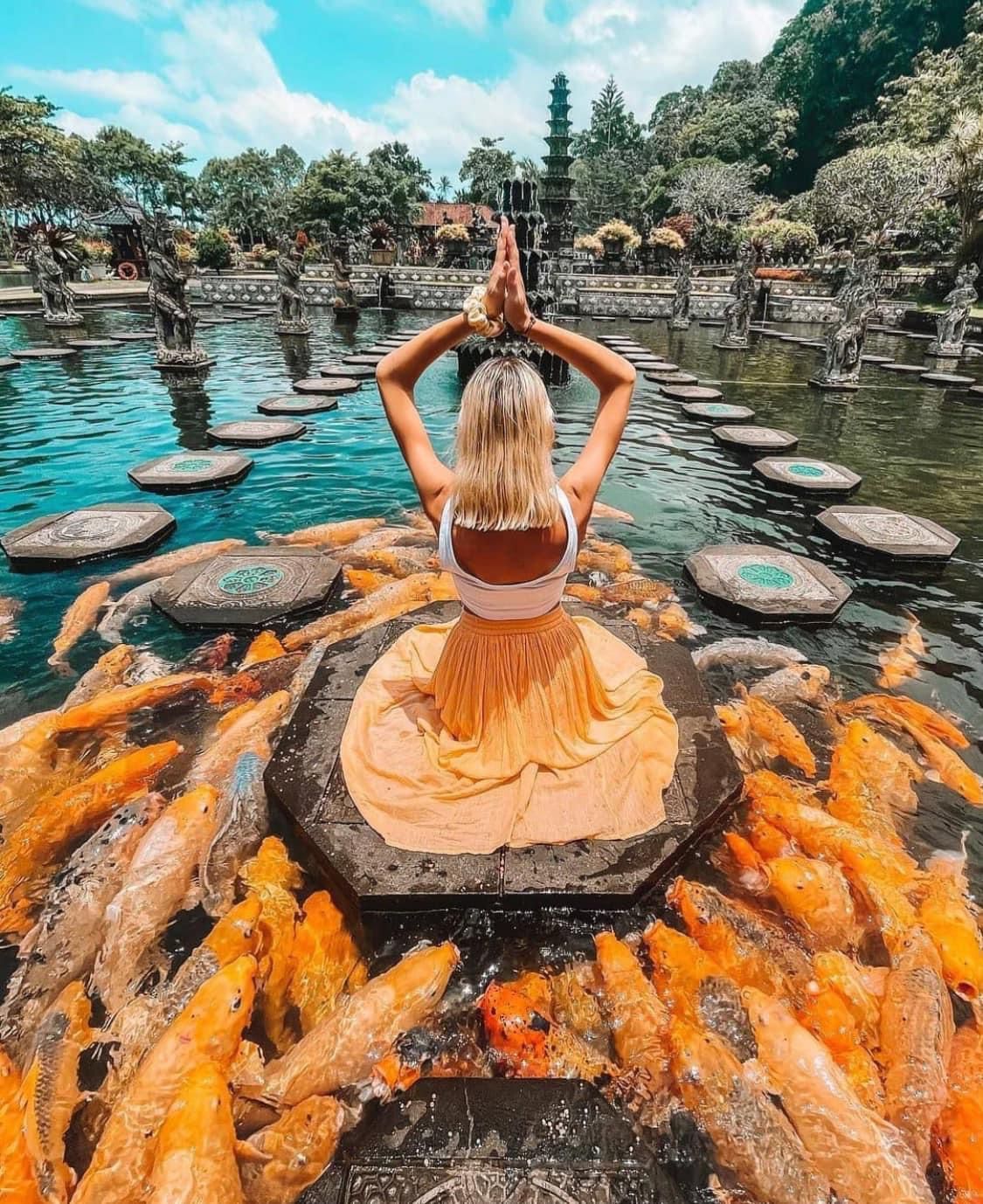 Woman in yellow skirt does yoga pose surrounded by orange koi fish in a pond.