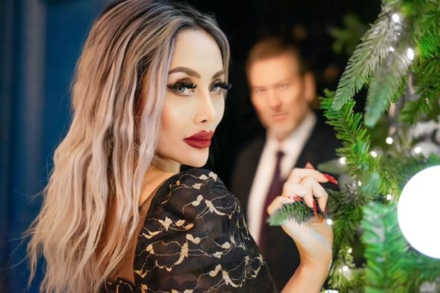 Woman with blonde hair and red lipstick, looking over her shoulder. Man in a suit in the background, Christmas tree nearby.