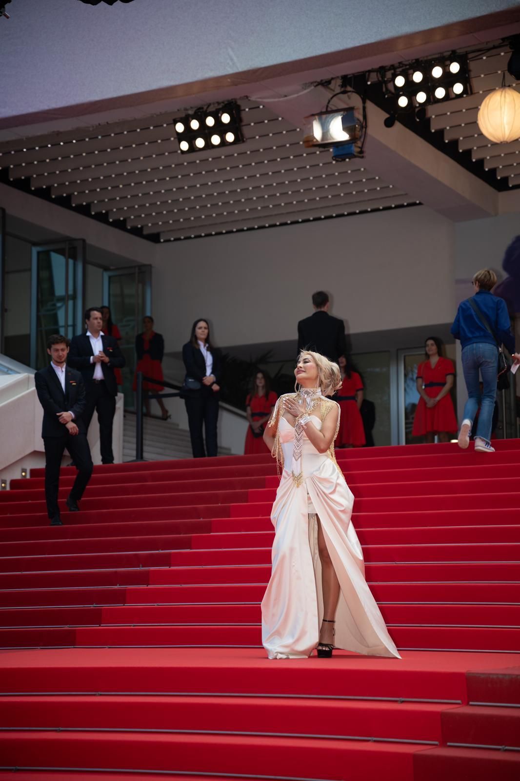 Woman in white gown on red carpet steps, posing for cameras. Cannes Film Festival backdrop.