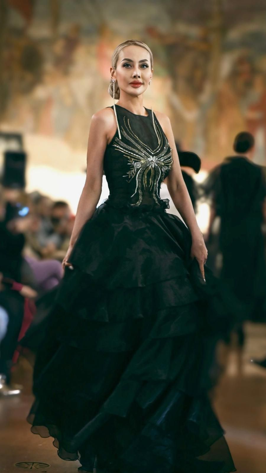 Woman in a black, tiered ball gown, embellished bodice, walking in a ballroom with a blurred background.