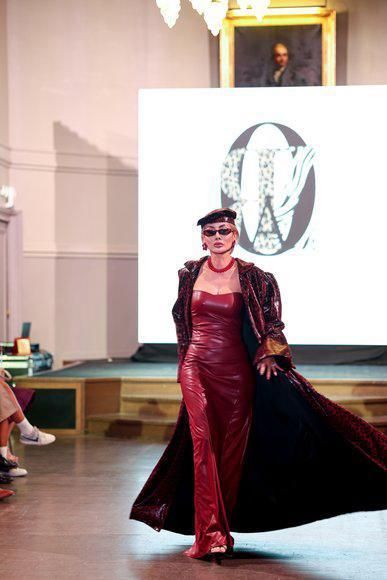 Woman in red gown and coat walks runway. Background: Logo, stage, audience, painting.