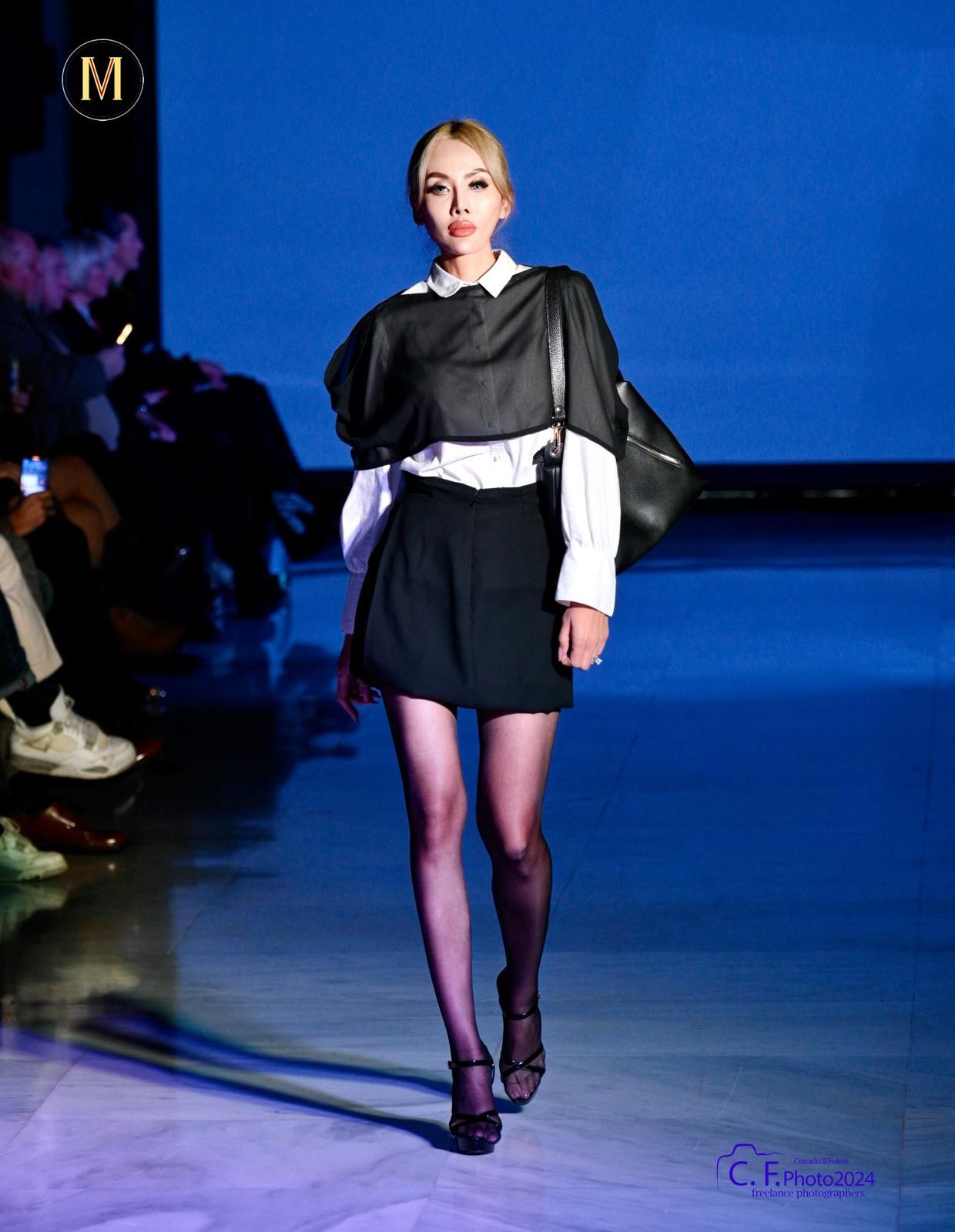 Model in black mini-skirt and top walks runway. White shirt and shoulder bag against a blue backdrop.