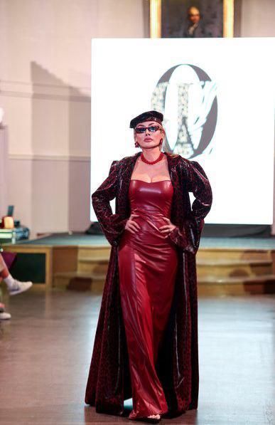 Woman in a red dress and coat walks a runway. She wears glasses and a hat. Backdrop features an 