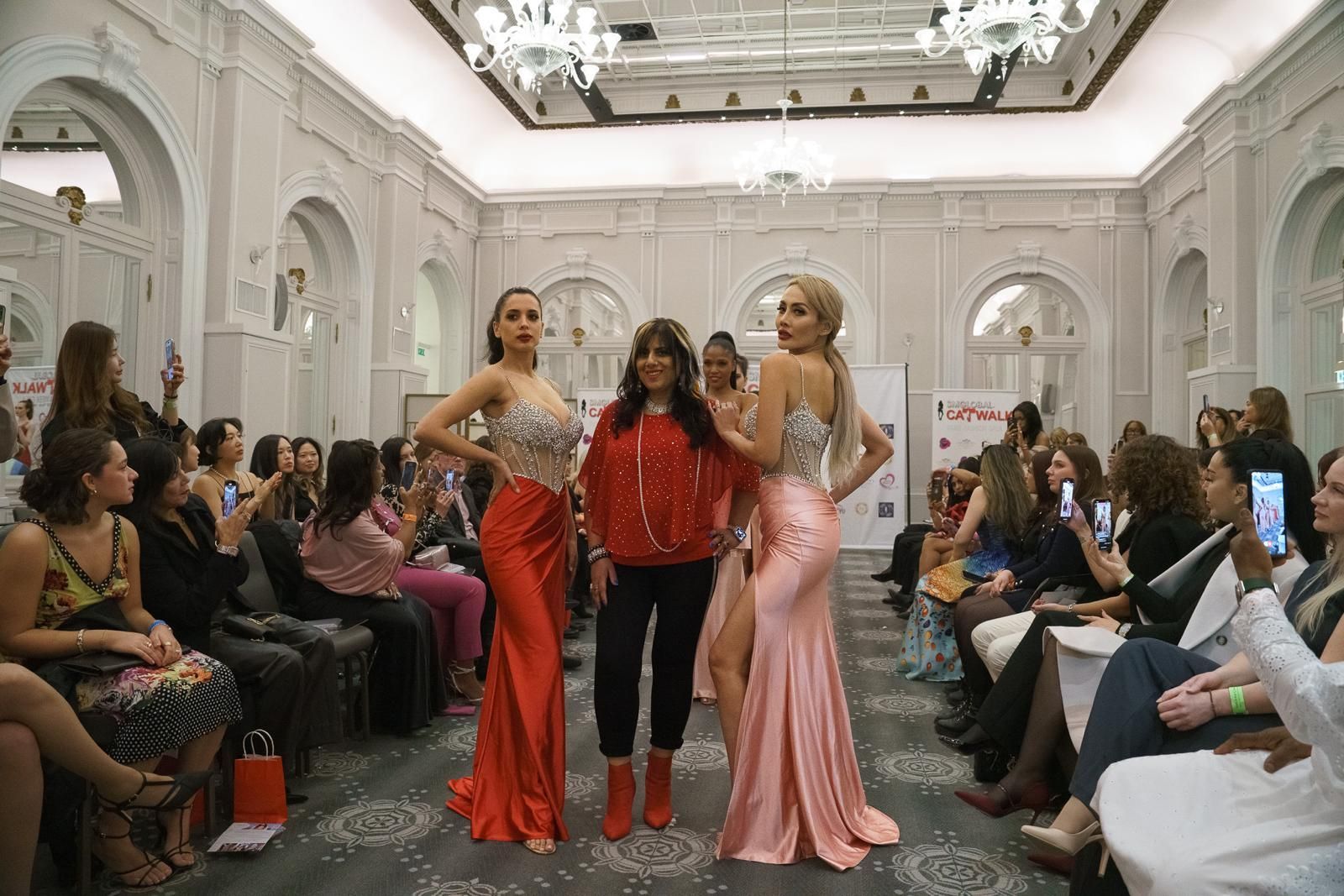 Fashion show: Models in gowns, designer in red jacket, posing on a runway. Audience seated.