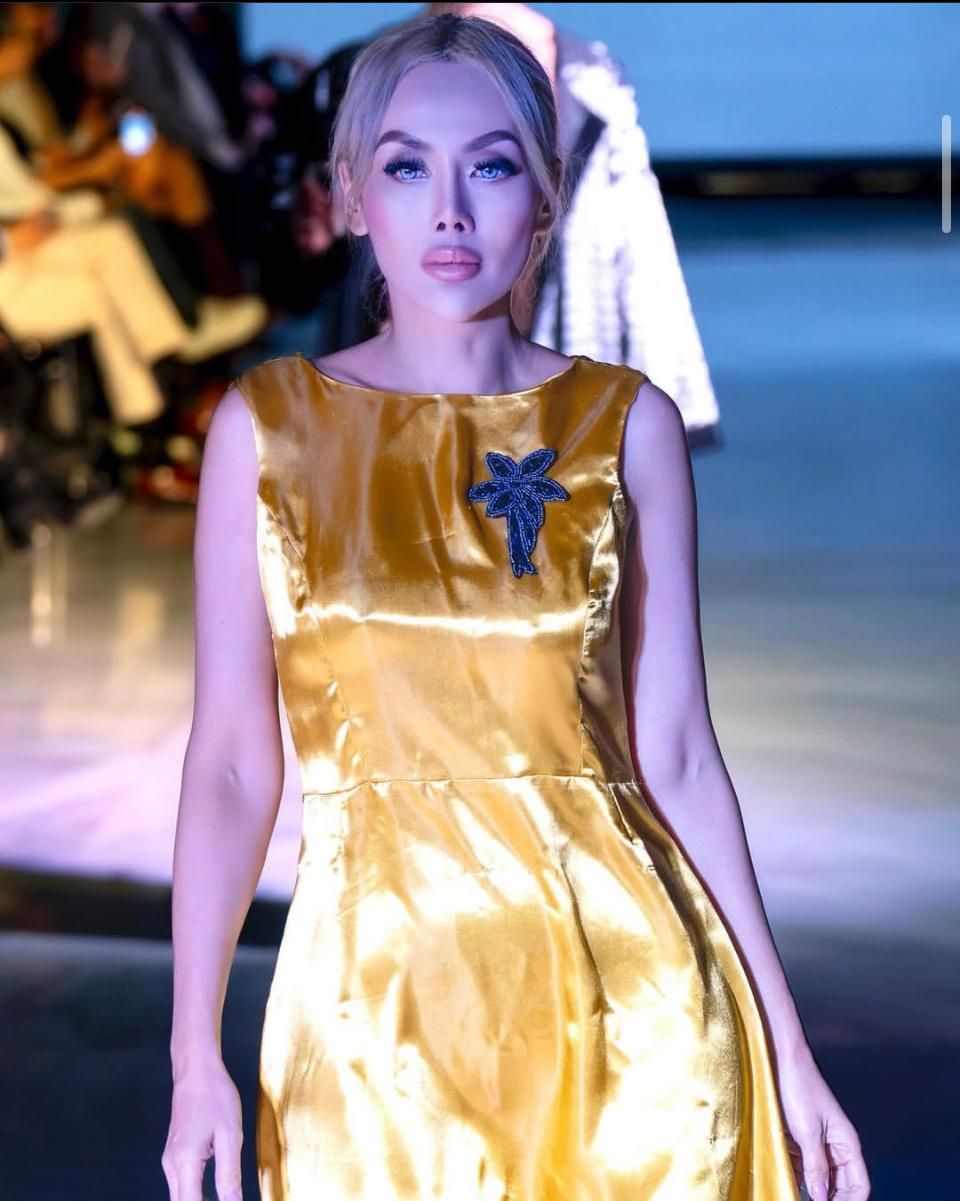 Woman modeling a gold satin dress with a dark blue brooch on a runway.