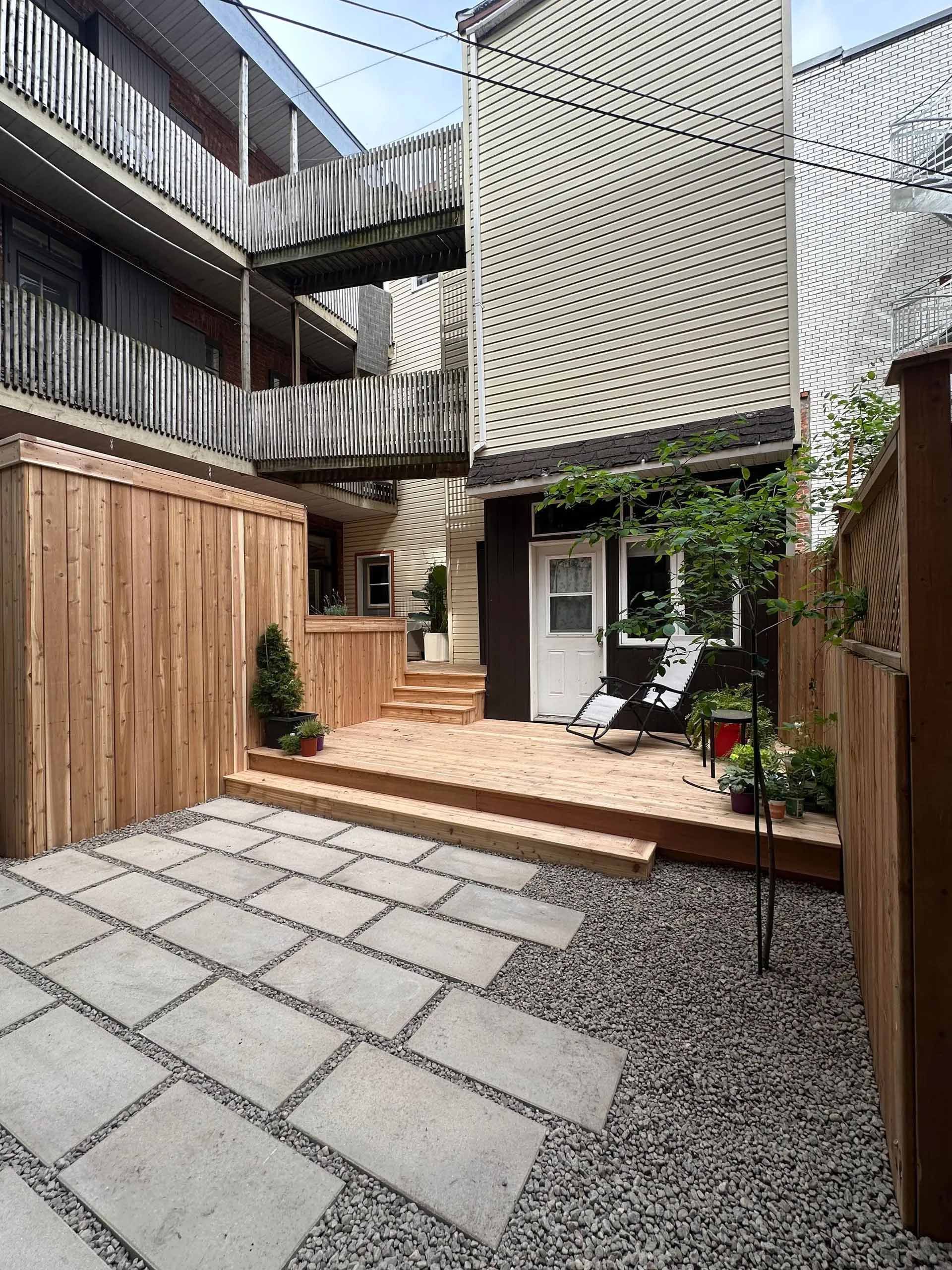 Petit jardin avec allée pavée, terrasse en bois et clôture en bois. Bâtiments en arrière-plan.