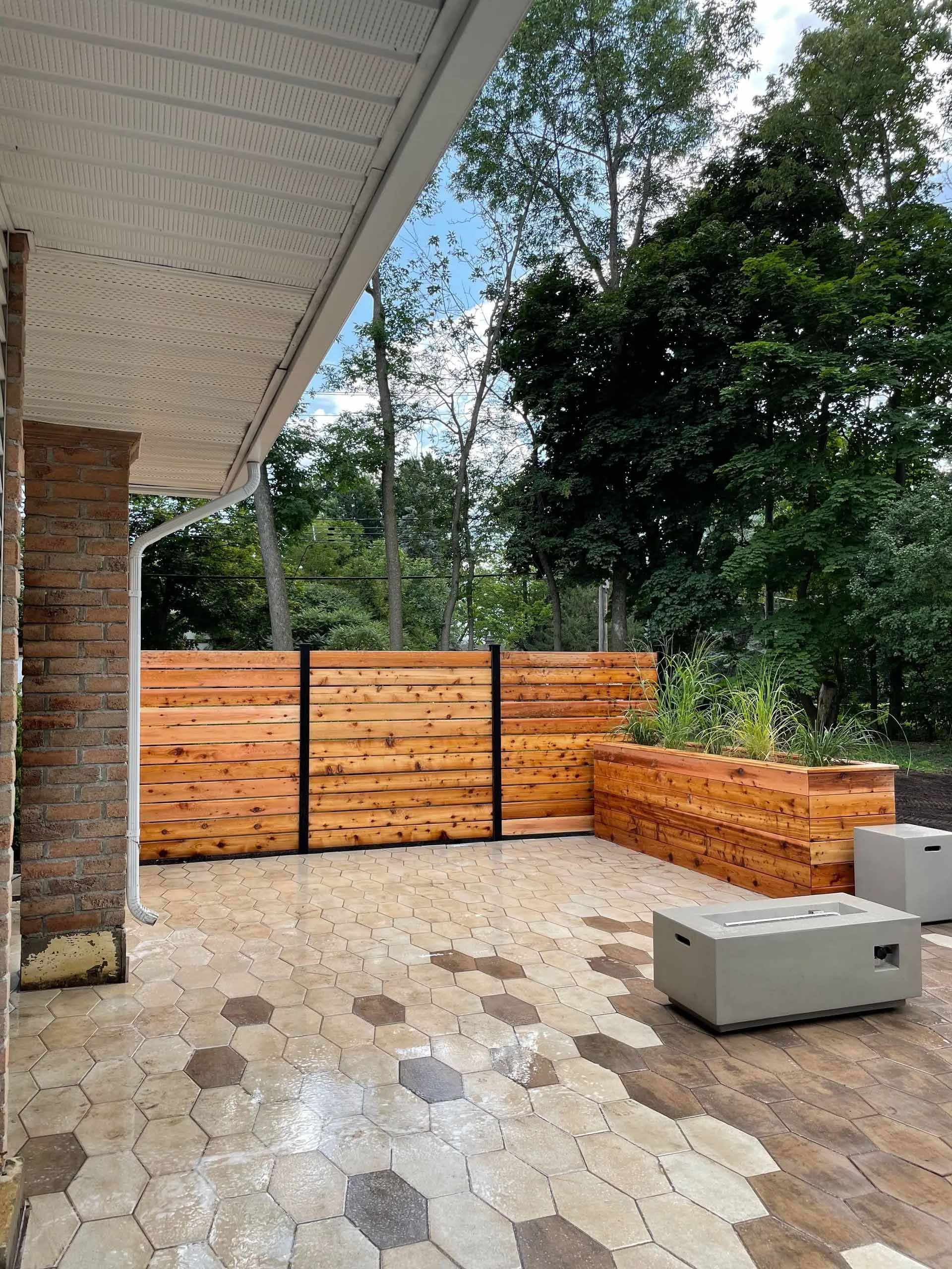 Terrasse avec clôture en bois et jardinières. Pavés bruns et gris, bâtiment beige. Arbres en arrière-plan.