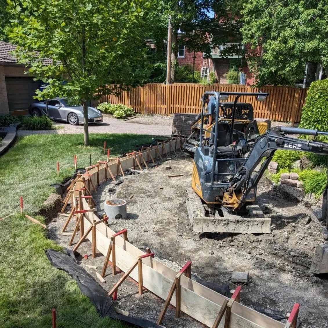 Chantier de construction avec une mini-pelle en train de façonner un coffrage en béton incurvé dans un jardin.