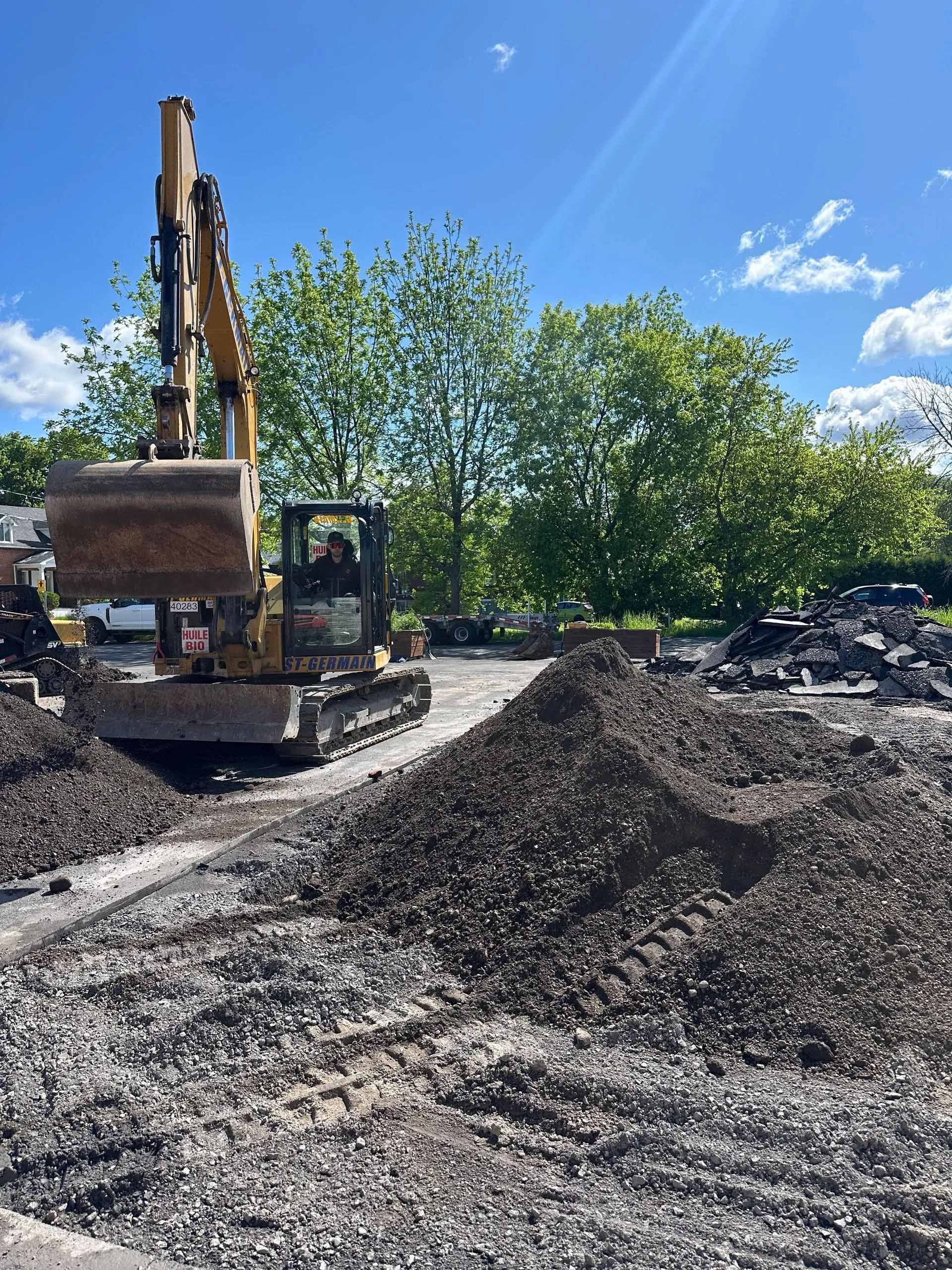 Une pelleteuse jaune travaille sur une surface de gravier sous un ciel bleu, avec des arbres en arrière-plan.