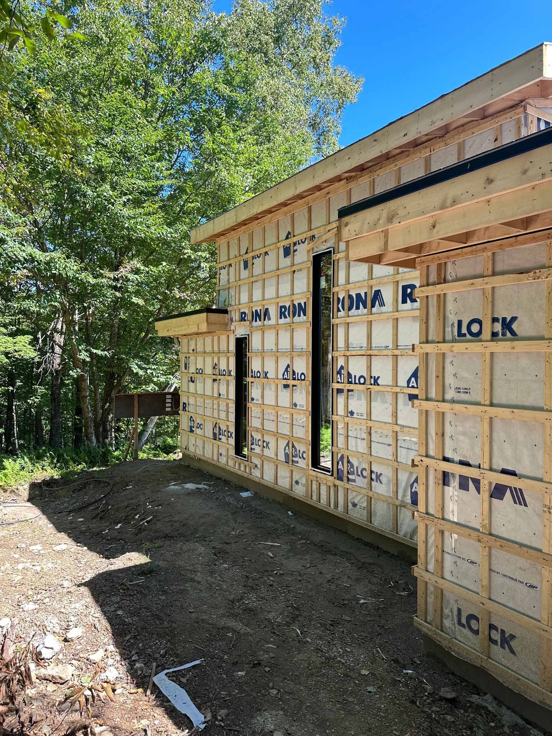 Extérieur d'un bâtiment en construction, montrant la charpente en bois, l'isolation et les ouvertures des fenêtres.